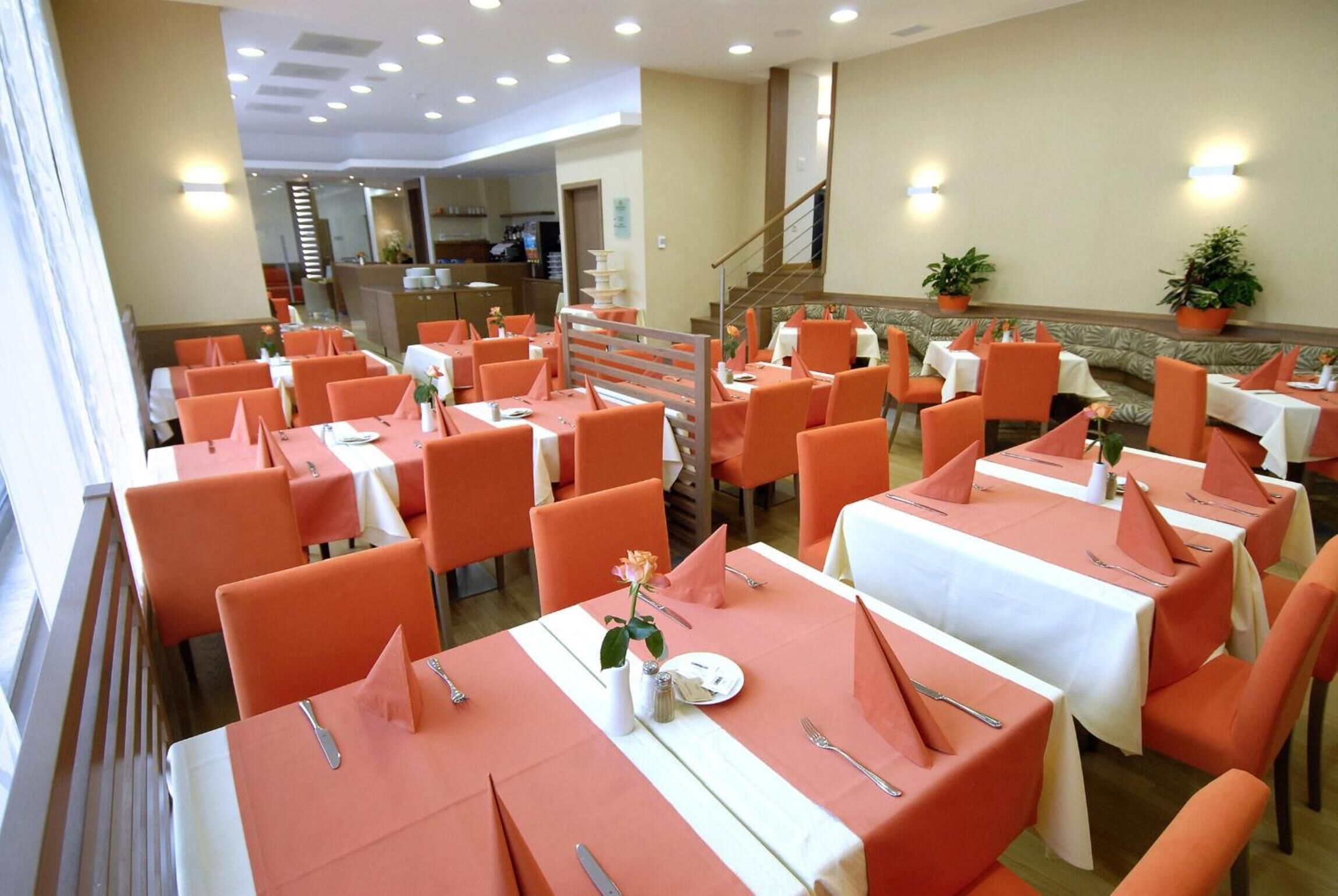 A dining area with tables set for four, orange chairs, and a staircase in the background.