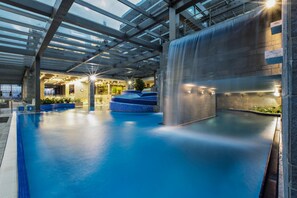 Une piscine intérieure agrémentée d’une cascade, entourée d’espaces de détente et dotée d’un toit en verre.