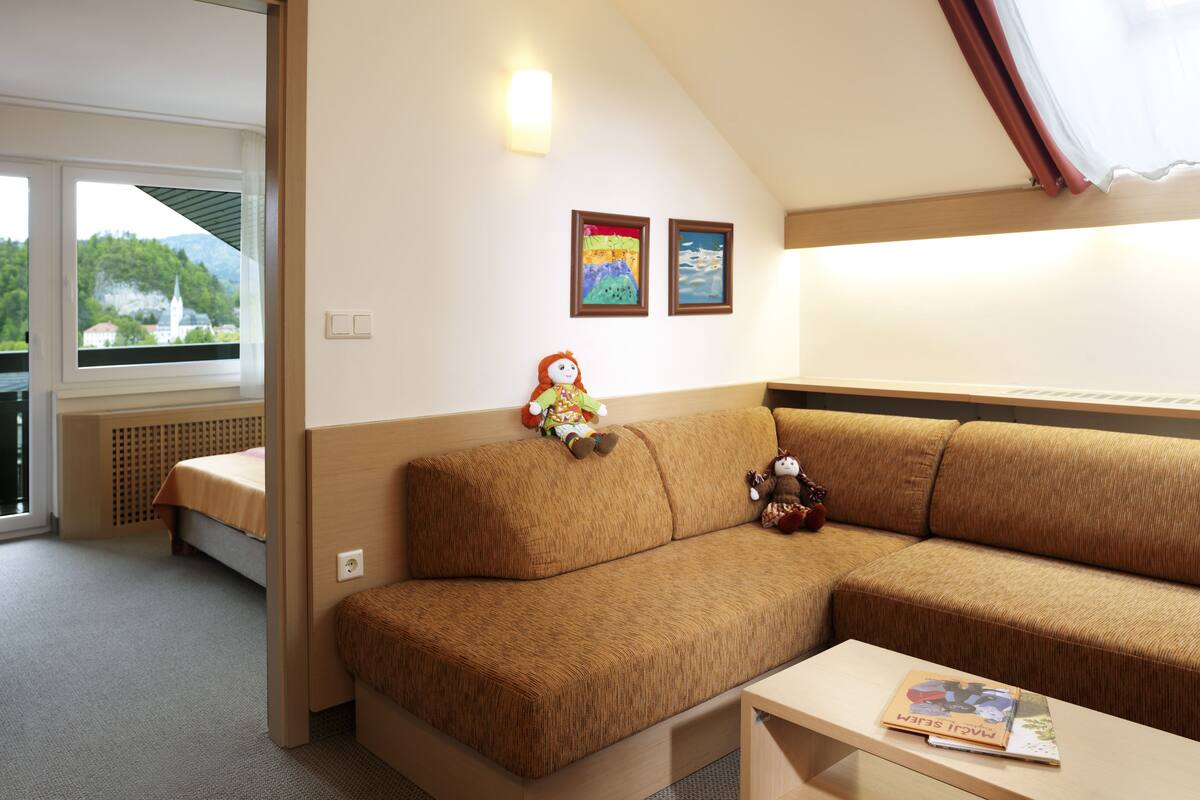 A cozy room with a brown sofa, a small table with a book, and a window with a view of greenery and a building.