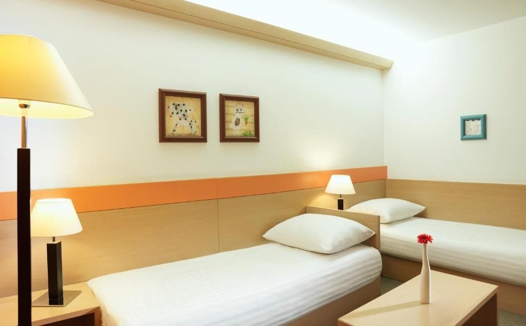 Ein Hotelzimmer mit zwei Einzelbetten, einem Nachttisch mit Lampe, einem kleinen Tisch mit Vase und zwei gerahmten Bildern an der Wand.