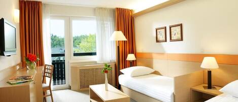 A hotel room with a bed, a desk, a chair, a small table, two bedside lamps, a TV, and a window with curtains.