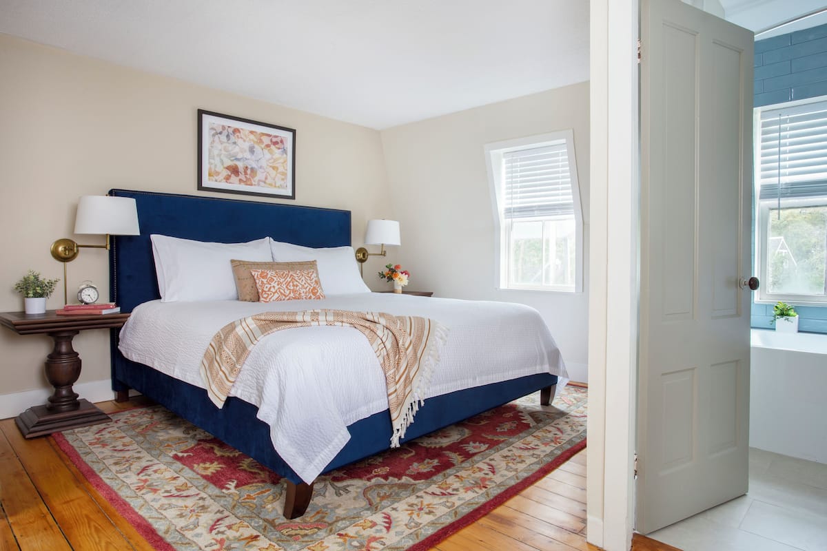 A bedroom with a large bed, two bedside tables, a nightstand, a window with blinds, and a door.