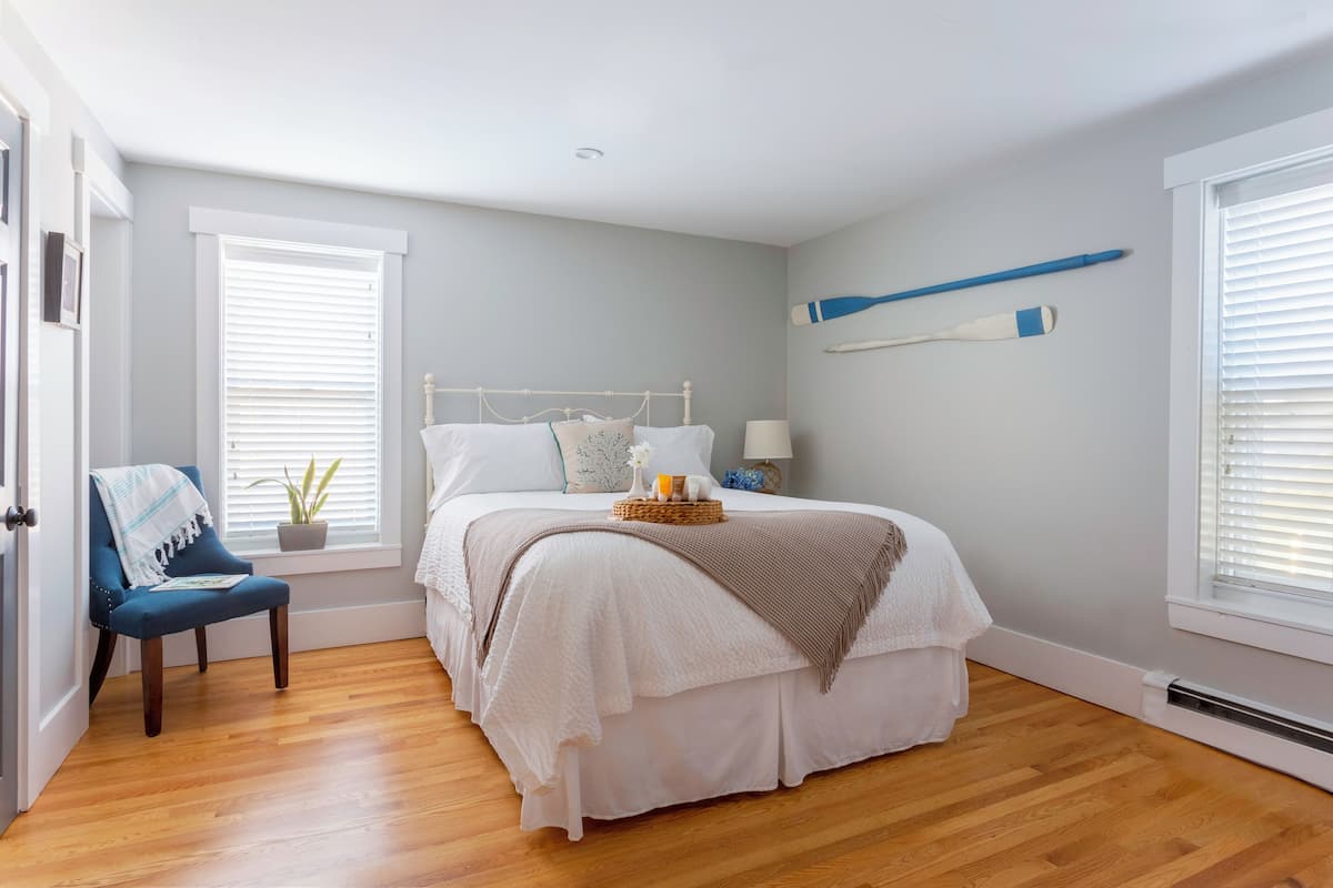 A bedroom with a bed, a chair, a bedside table, a lamp, and a window with blinds.
