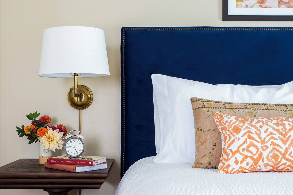 A neatly made bed with a patterned blanket, a bedside table with a lamp, a clock, and a vase with flowers.
