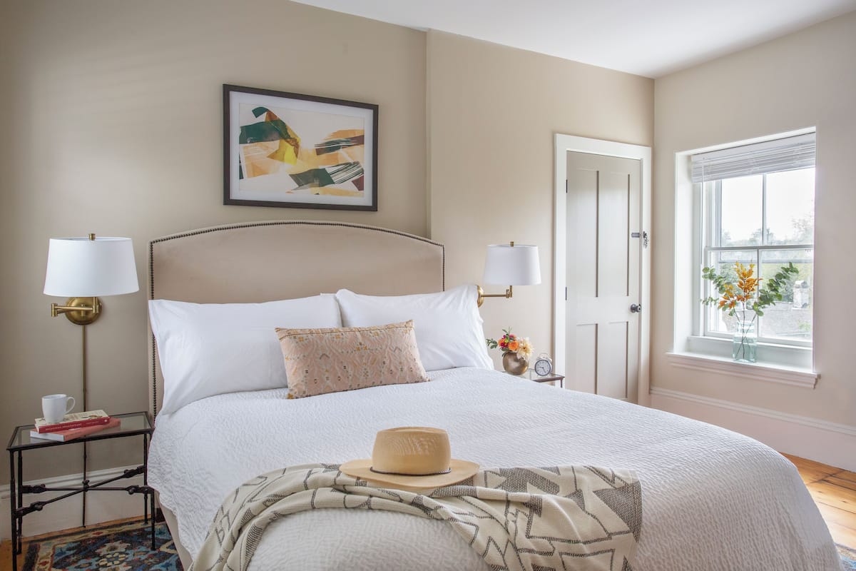A neatly arranged bedroom with a bed, bedside lamps, a framed artwork, a window with a view, and a bedside table with books and a cup.