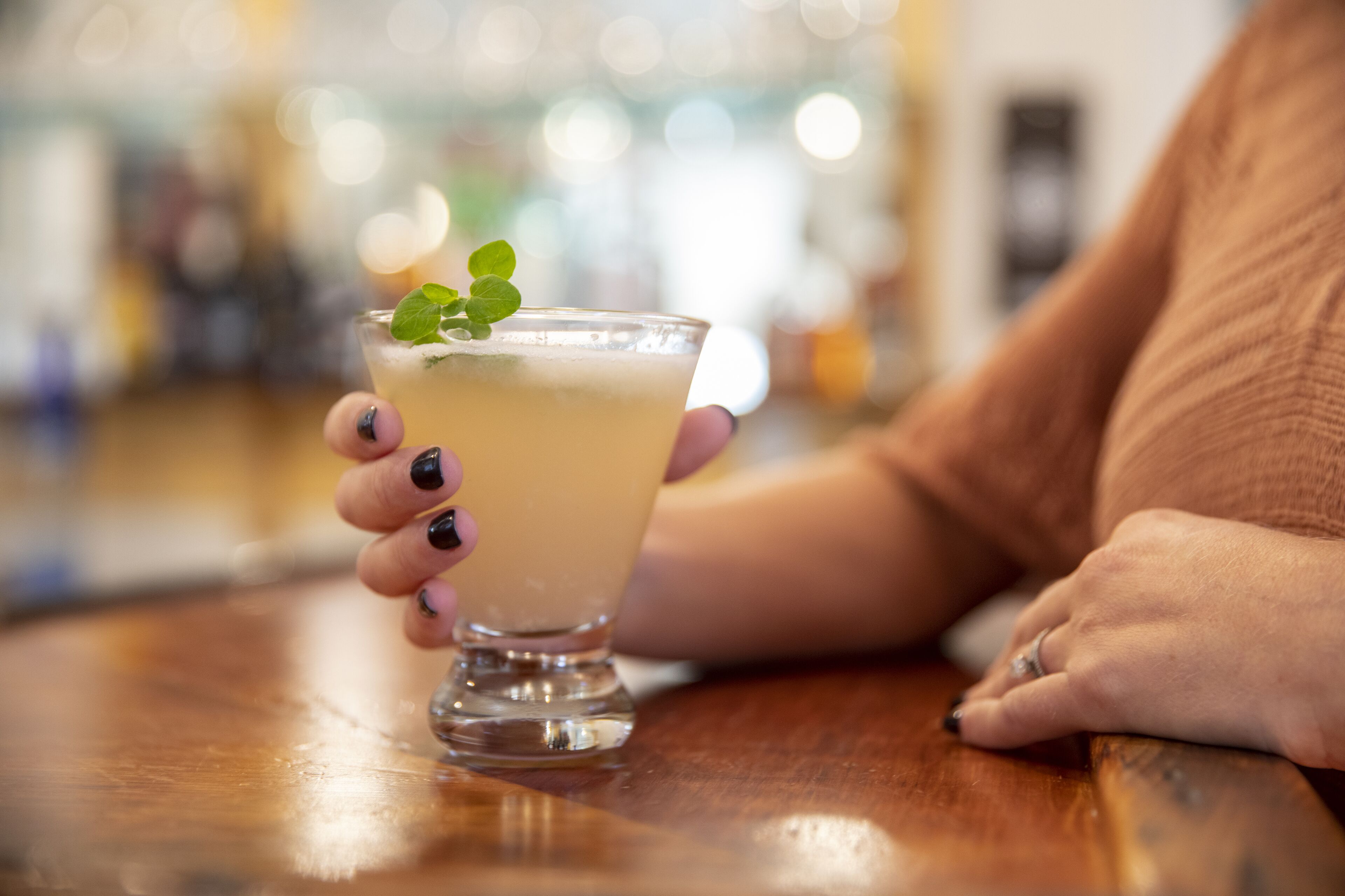 Eine Person hält ein Glas mit einem hellen Getränk und einer Minze, sitzt an einem Holztisch in einem gut beleuchteten Innenraum.