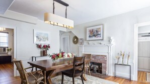 A dining room with a wooden table, wicker chairs, a chandelier, a fireplace, and a floral arrangement. - Chapter House (Yarmouth Port)