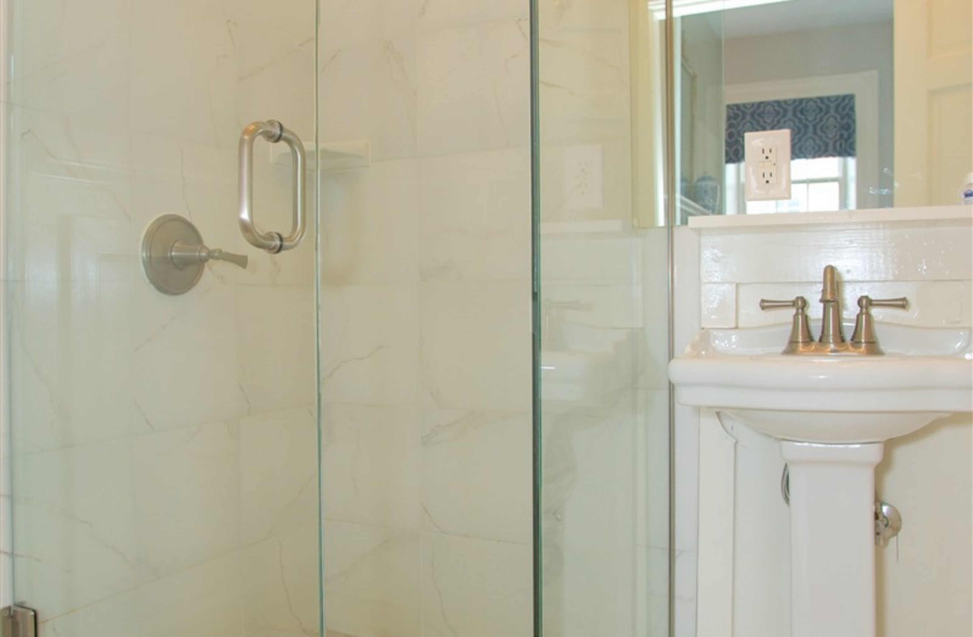 Une salle de bain dotée d’une porte de douche en verre, d’un lavabo blanc avec des robinets dorés et d’un miroir qui reflète une fenêtre à motifs.