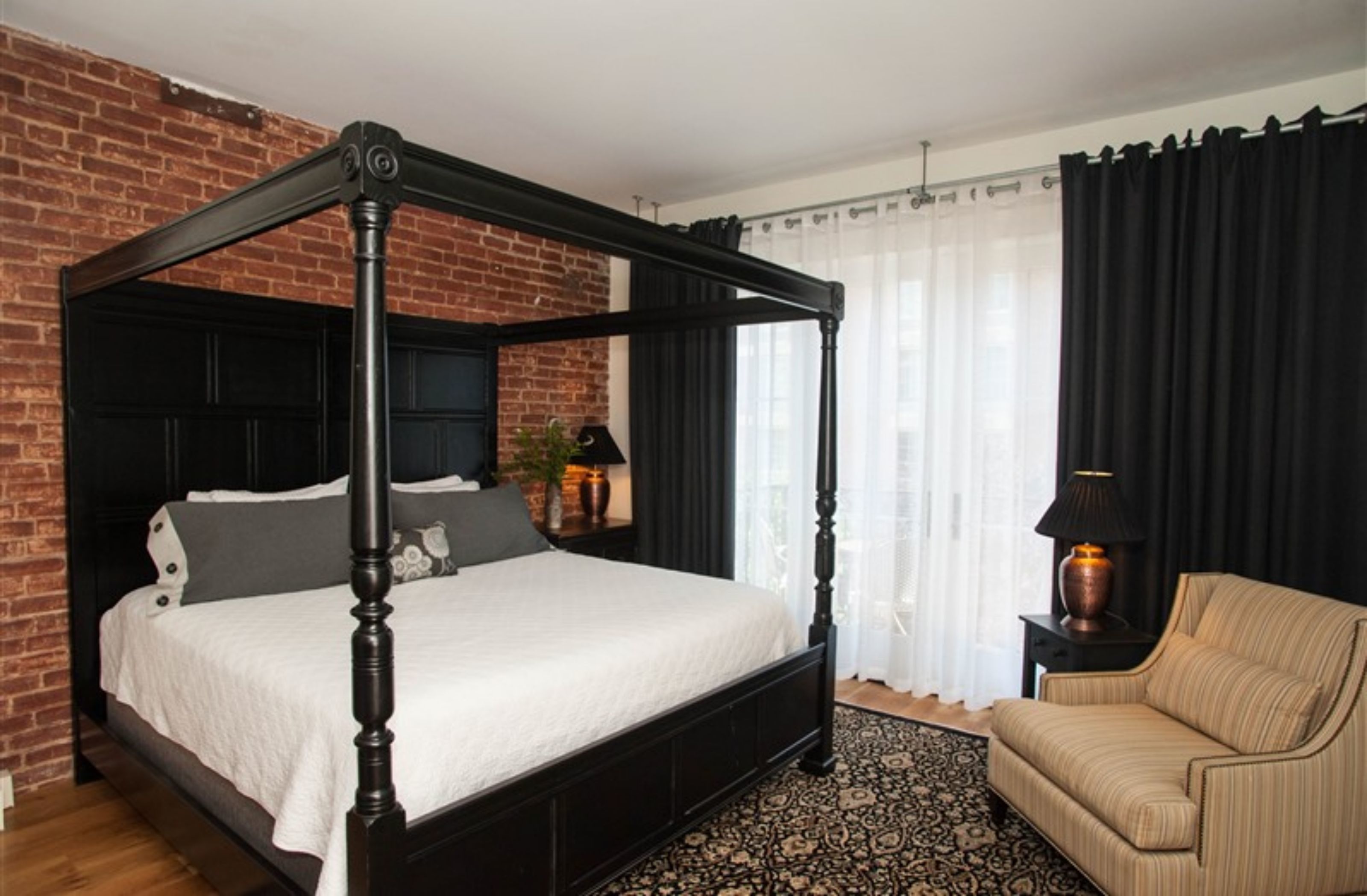 Un lit à baldaquin dans une pièce avec un mur en briques, un rideau noir et un tapis à motifs.