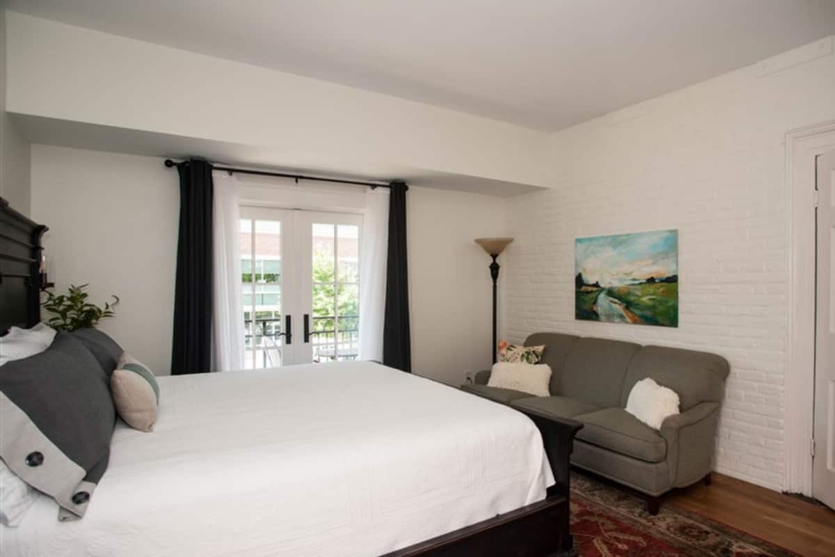 A bedroom with a large bed, a grey sofa, a painting on the wall, and a view of the outdoors through a window.