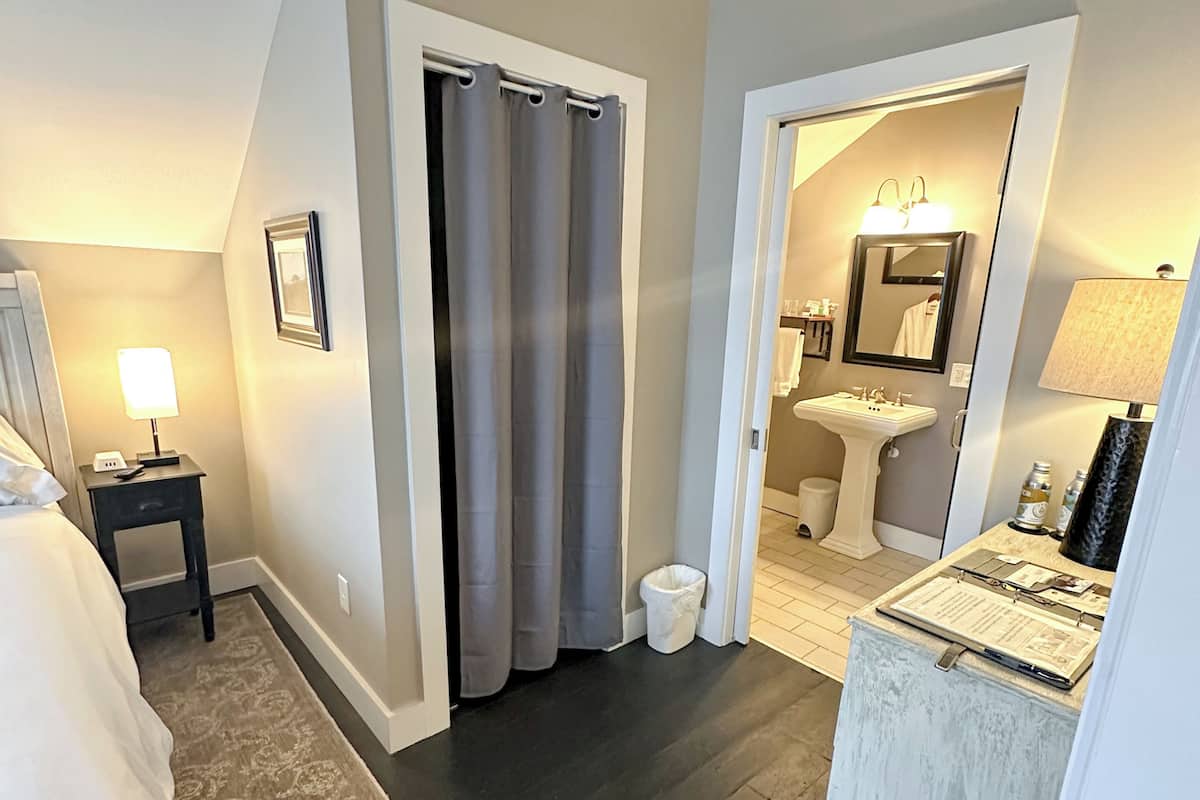 A hotel room with a bed, a nightstand, a desk with a lamp, a bathroom with a sink and mirror, and a shower area.