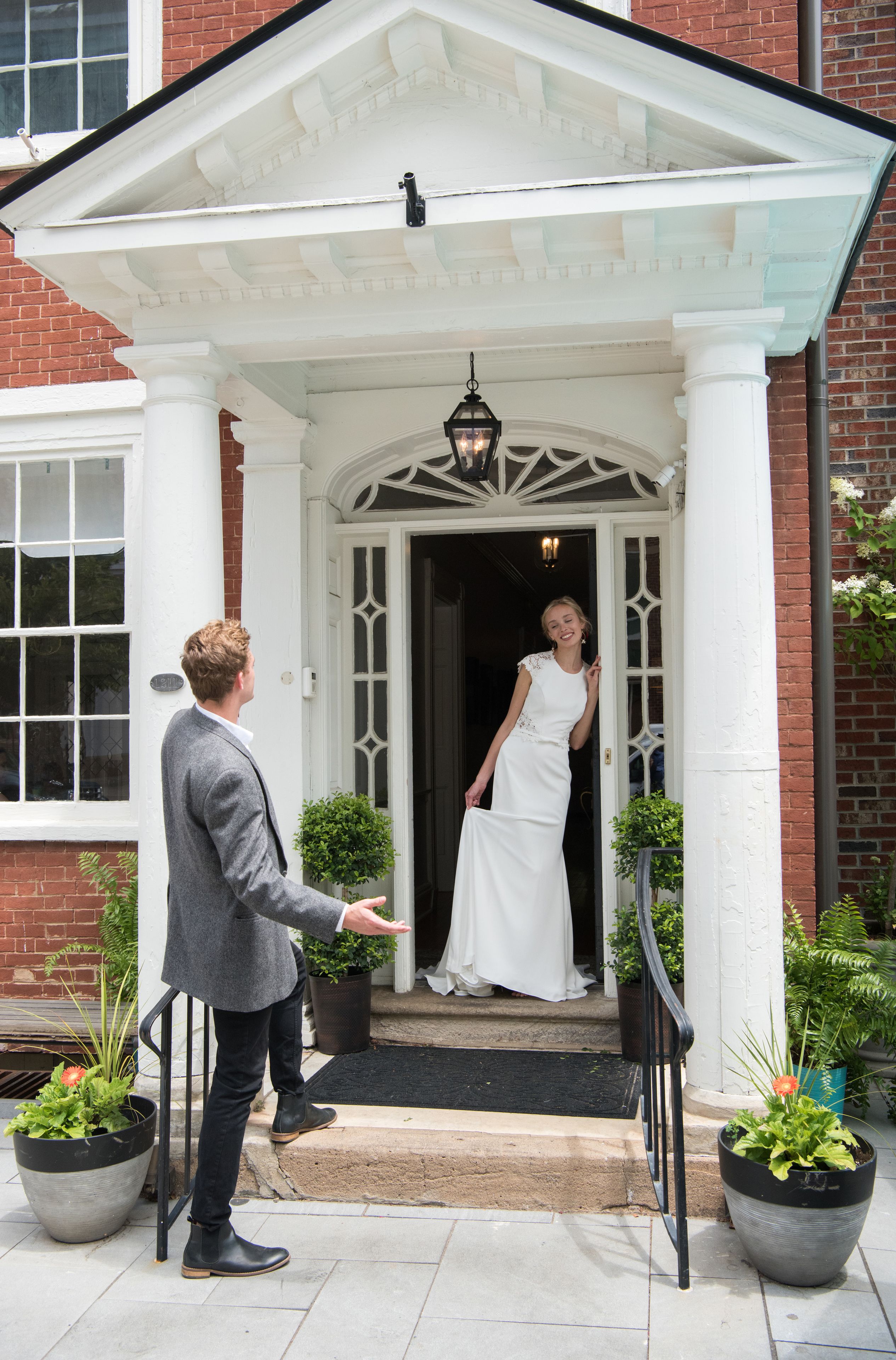 A man and a woman at a building entrance with a white porch, a black metal railing, and potted plants.