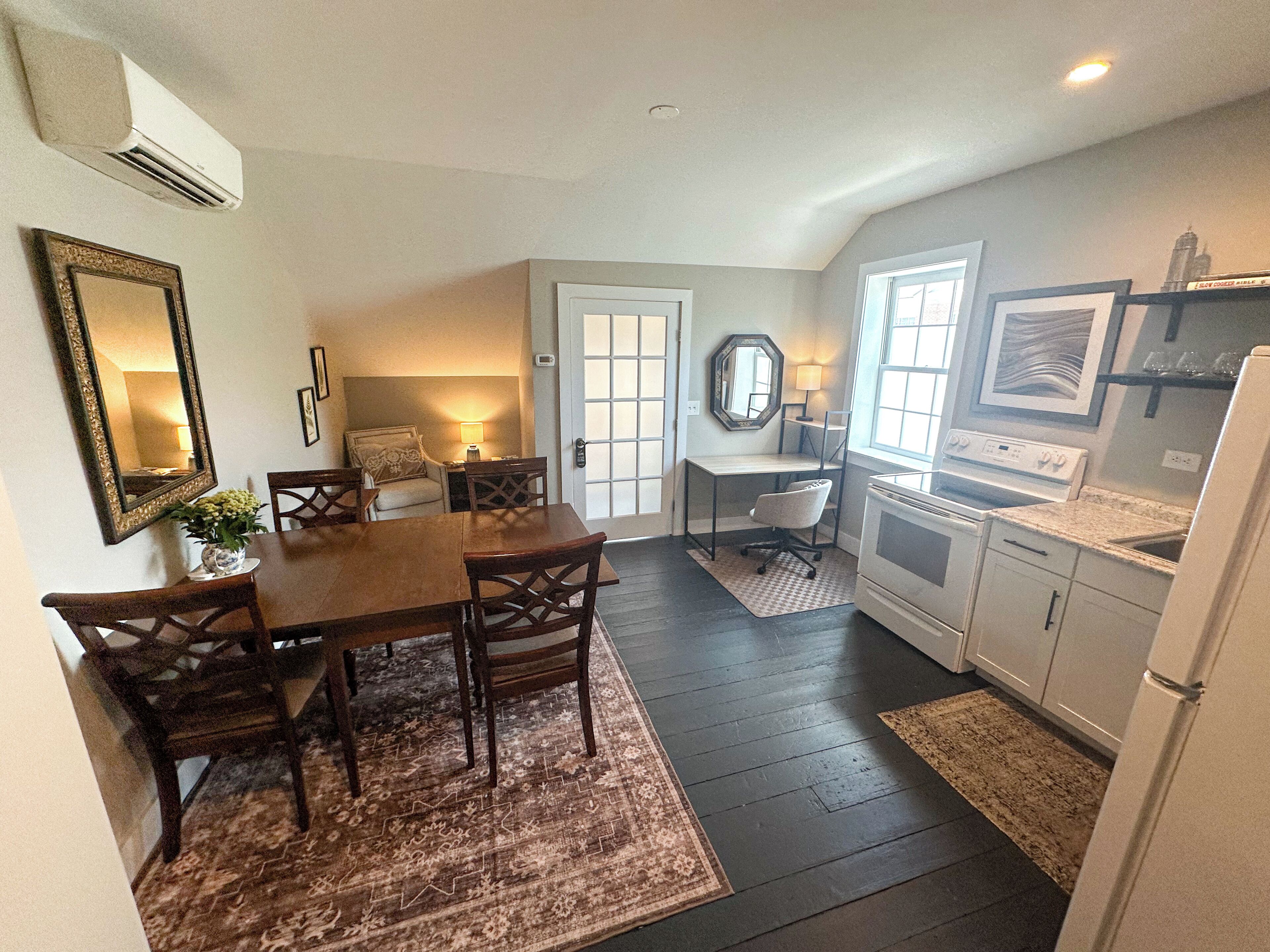 A dining area with a wooden table and chairs, a kitchen with a stove and refrigerator, and a door leading to another room.
