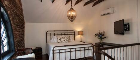 Une chambre à coucher comprenant un lit, un luminaire suspendu, une table de chevet, une chaise, une fenêtre et une rambarde d’escalier.