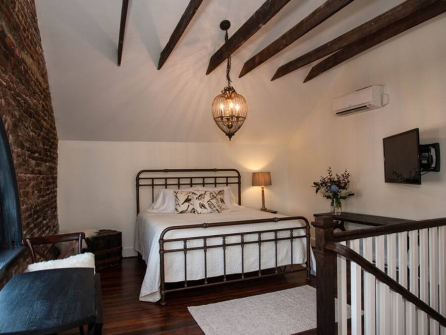 Une chambre à coucher comprenant un lit, un luminaire suspendu, une table de chevet, une chaise, une fenêtre et une rambarde d’escalier.