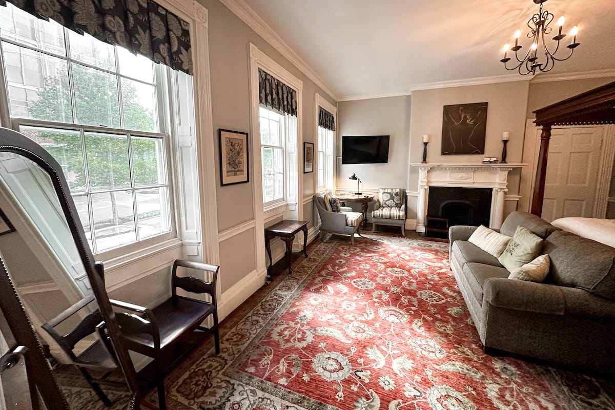 A living room with a fireplace, a sofa, a dining table with chairs, and a large window with curtains.