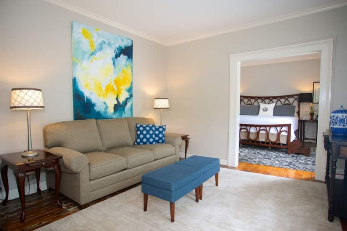 A living room with a beige sofa, a blue ottoman, and a large abstract painting on the wall.