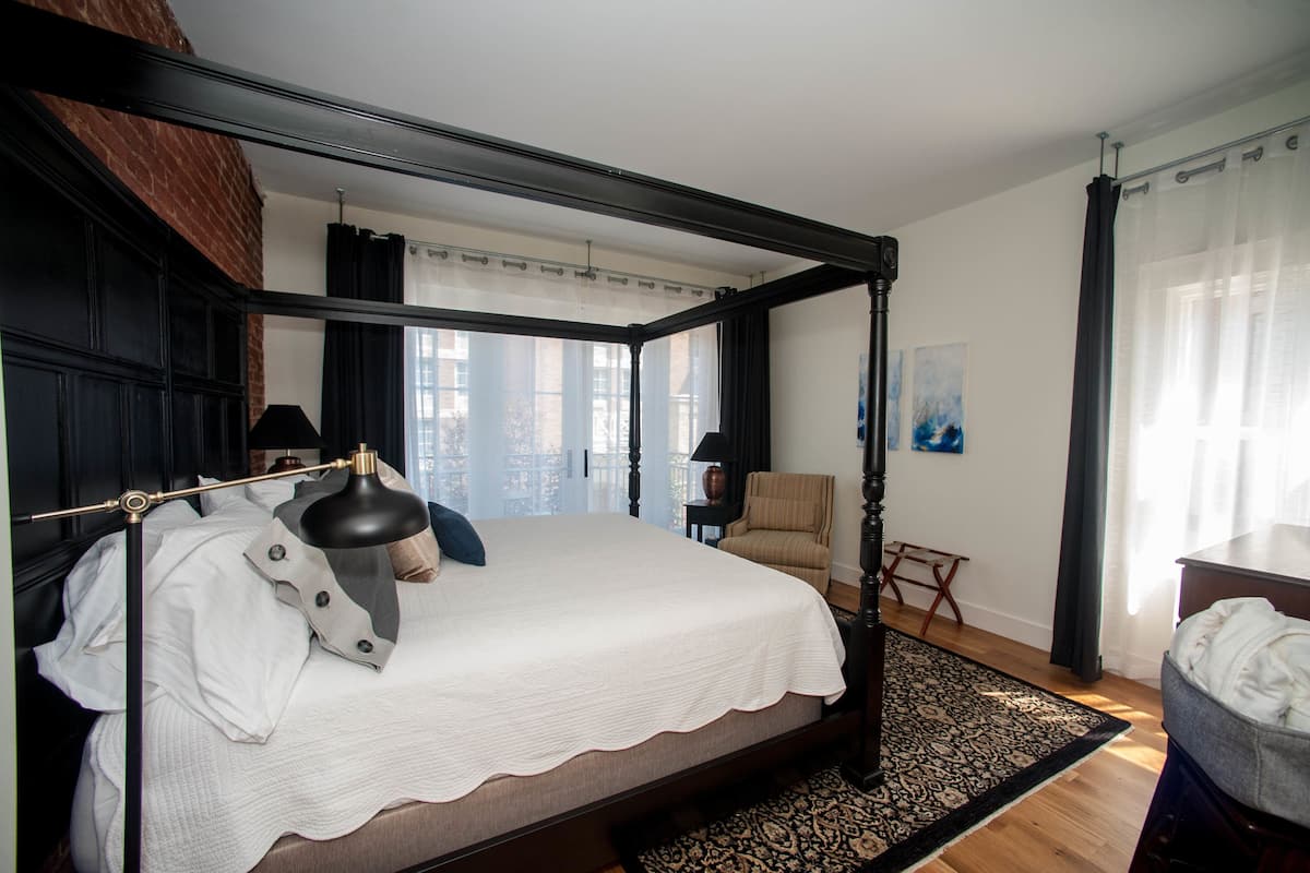 A four-poster bed with a white bedspread, a bedside lamp, and a chair in a room with a large window and a view of buildings.