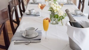 A dining area with a long table set for a meal, including a bottle of champagne, a glass of orange juice, and a vase of flowers. - Dinsmore Boutique Inn (Charlottesville)