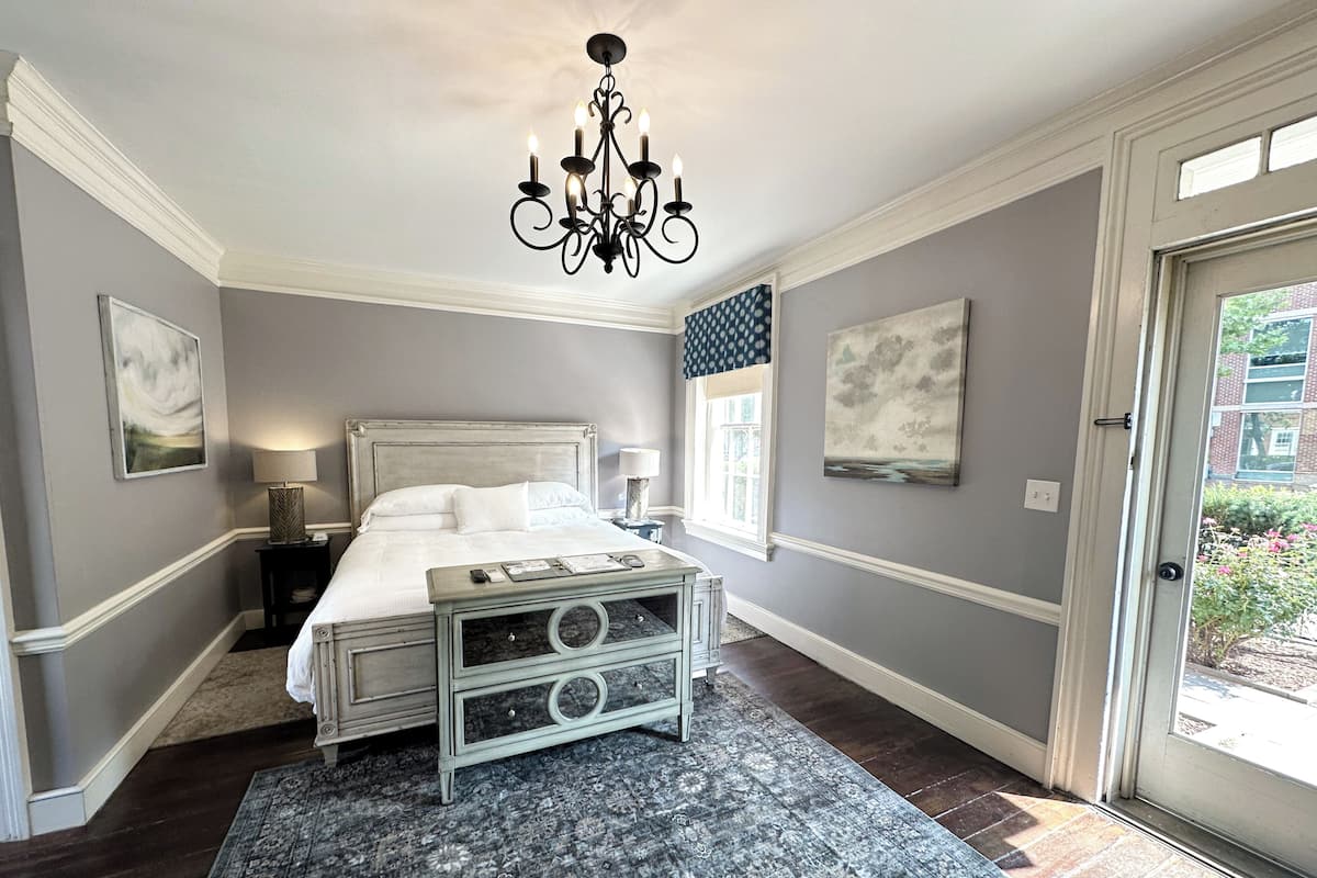 A bedroom with a bed, a chandelier, a nightstand, a dresser, a window, and artwork on the walls.