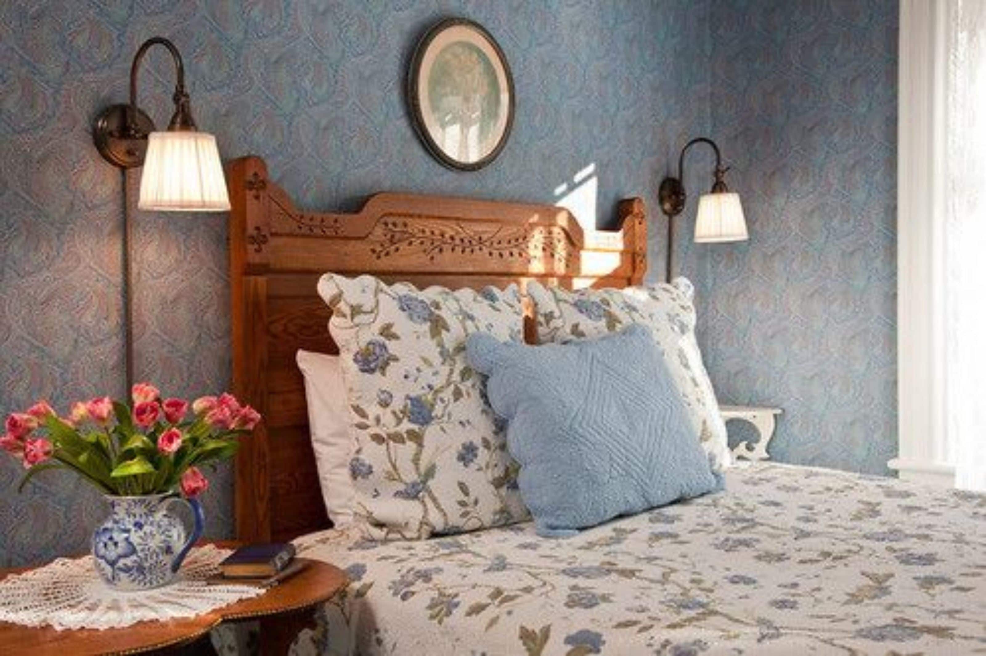 Une chambre à coucher avec une tête de lit en bois, une literie à motifs floraux, un vase de fleurs et des lampes fixées au mur.