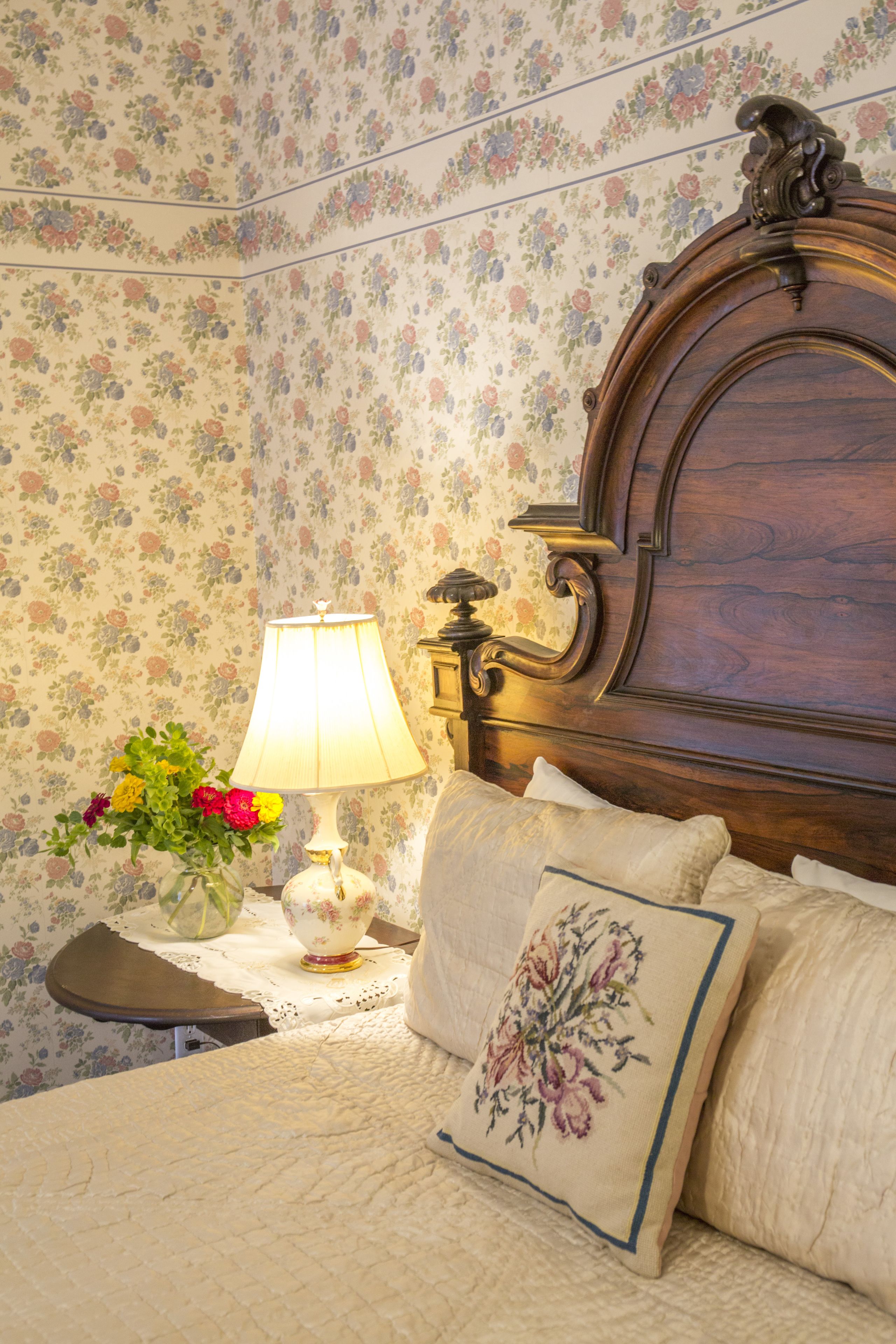 A neatly made bed with floral pillows, a bedside table with a lamp and a vase of flowers, and a wooden headboard.