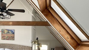 Een slaapkamer met kap en een plafondventilator, een vloerlamp, een bed met een bedspread met patroon, een stoel en een kleine tafel.