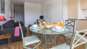 Un salon moderne avec une table à manger en verre, préparée pour le petit-déjeuner, un canapé et un téléviseur en arrière-plan.