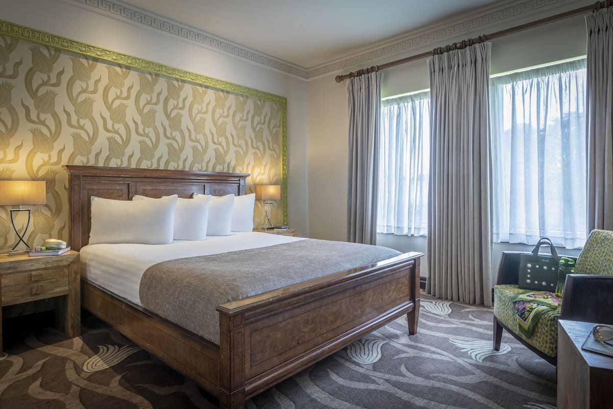 Habitación de hotel con una cama grande, dos lámparas de noche, un cabecero de madera, alfombra estampada, ventana con cortinas y una silla con cojín floral.