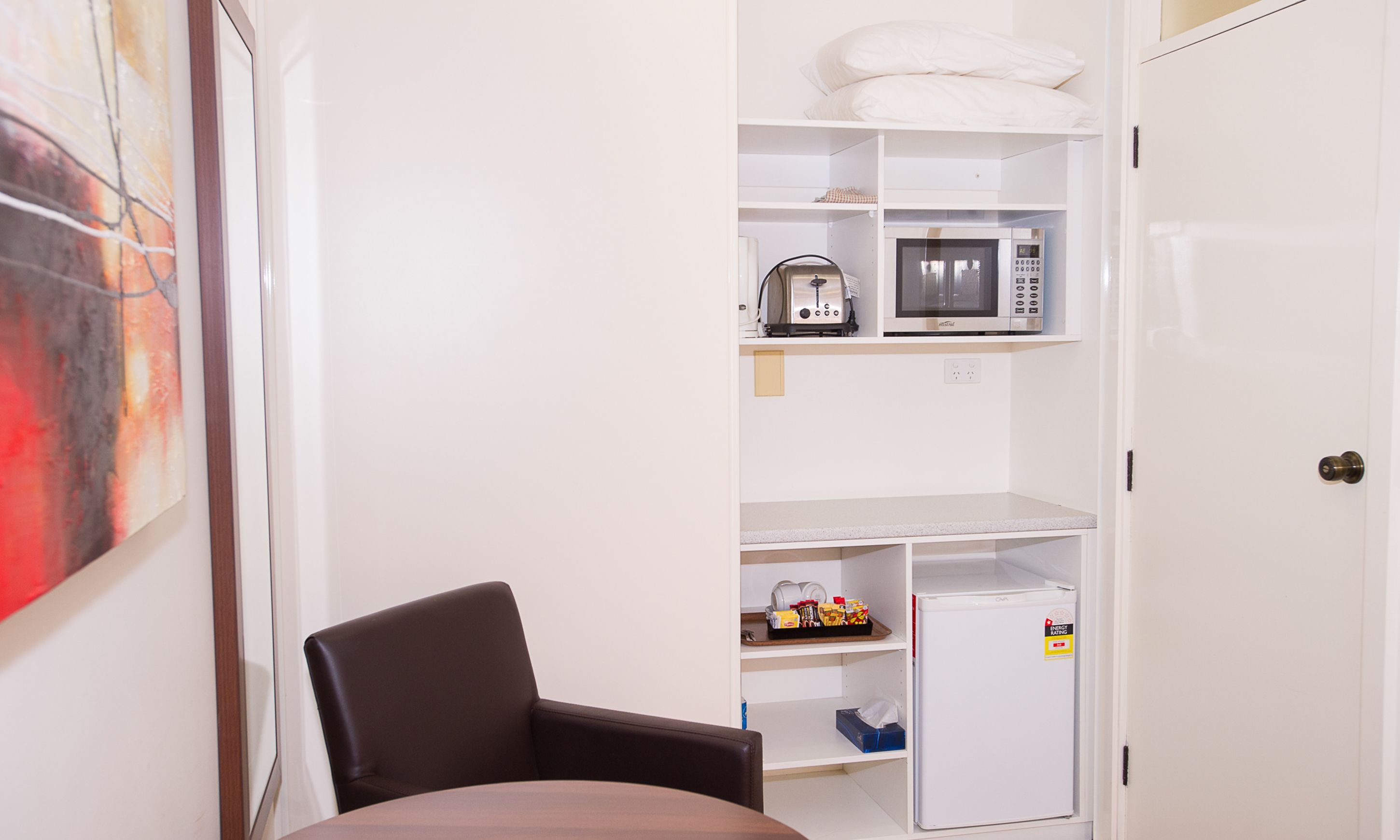 A compact room with a dining table, a chair, a microwave, a refrigerator, and a shelf with various items.