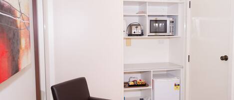 A compact room with a dining table, a chair, a microwave, a refrigerator, and a shelf with various items.