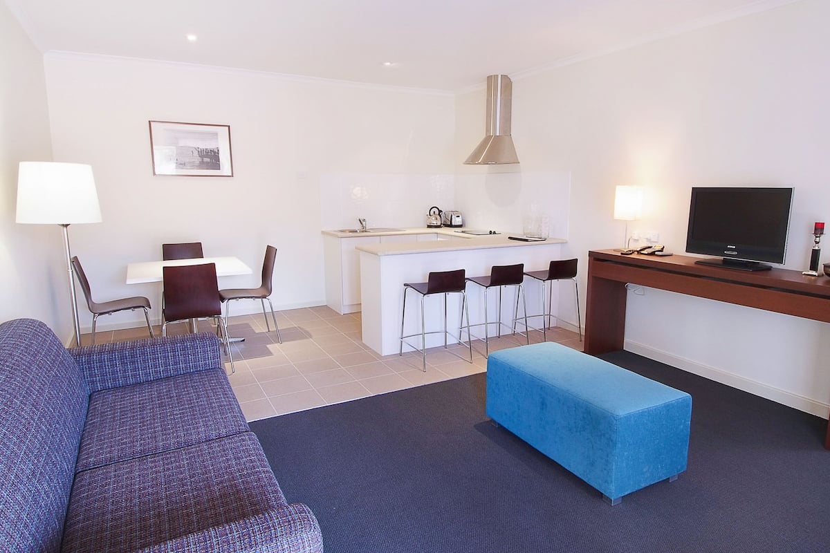 Uma sala de estar moderna com sofá, um banquinho azul, área de jantar com cadeiras, cozinha com balcão e uma TV sobre uma mesa de madeira.