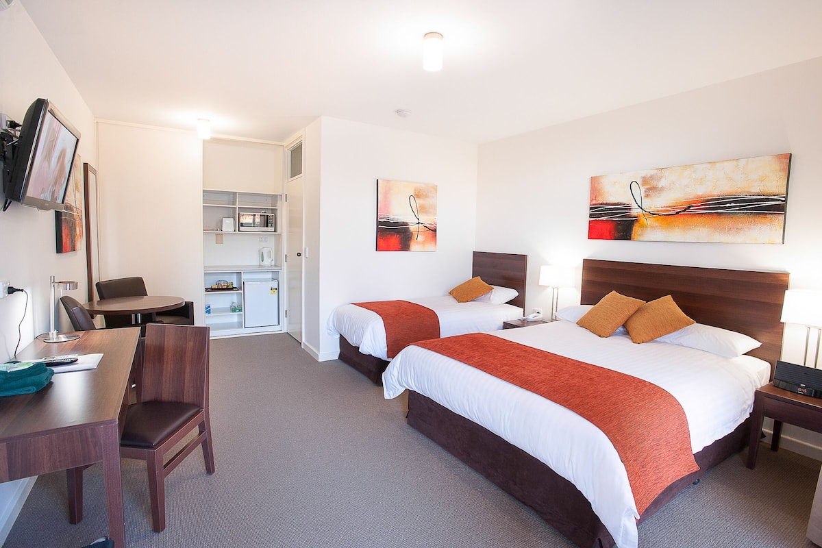 Quarto de hotel com duas camas, uma mesa, uma cadeira e uma cozinha compacta.