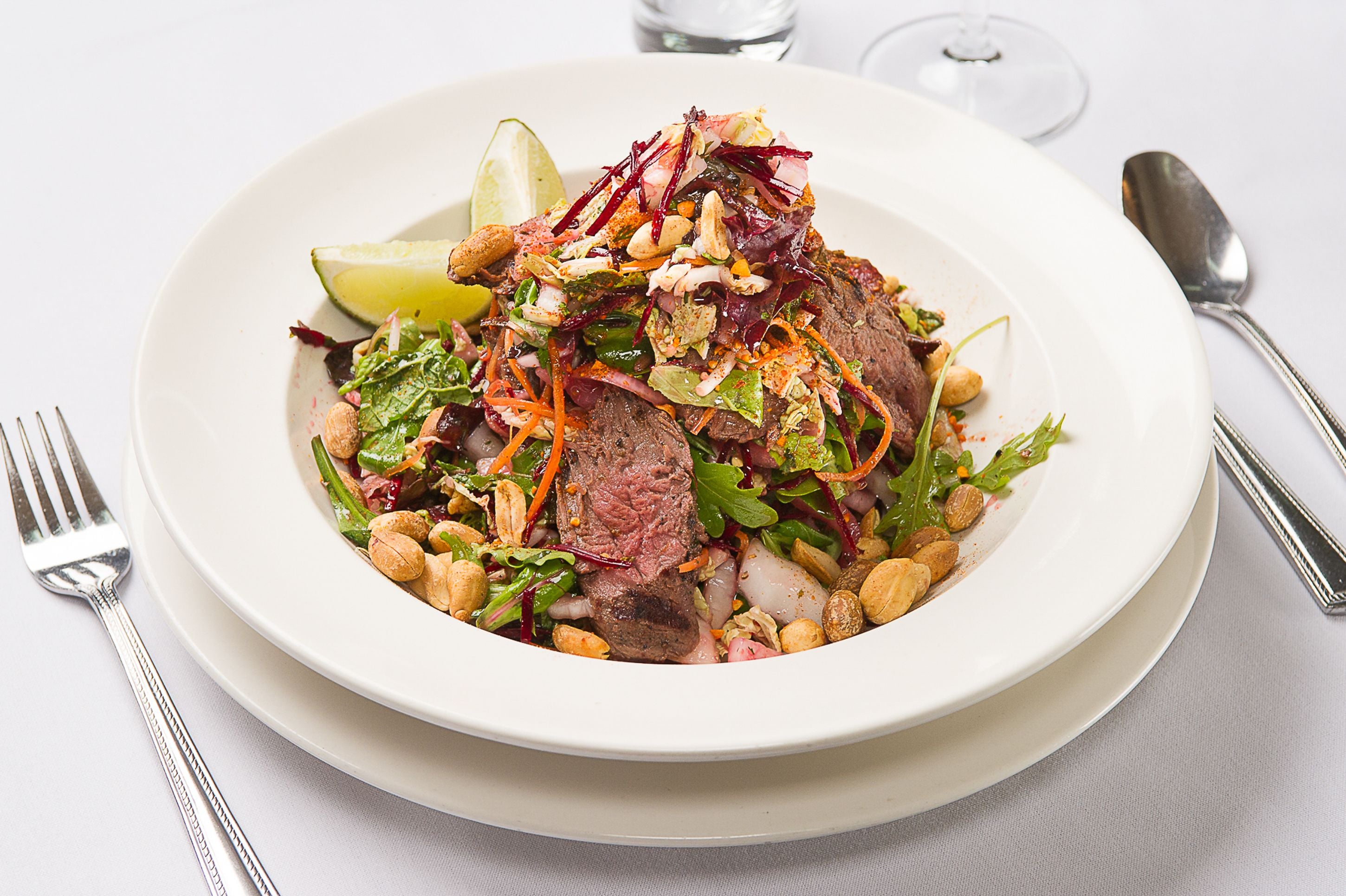 A white plate with a salad and steak, accompanied by a fork and spoon on a white tablecloth.
