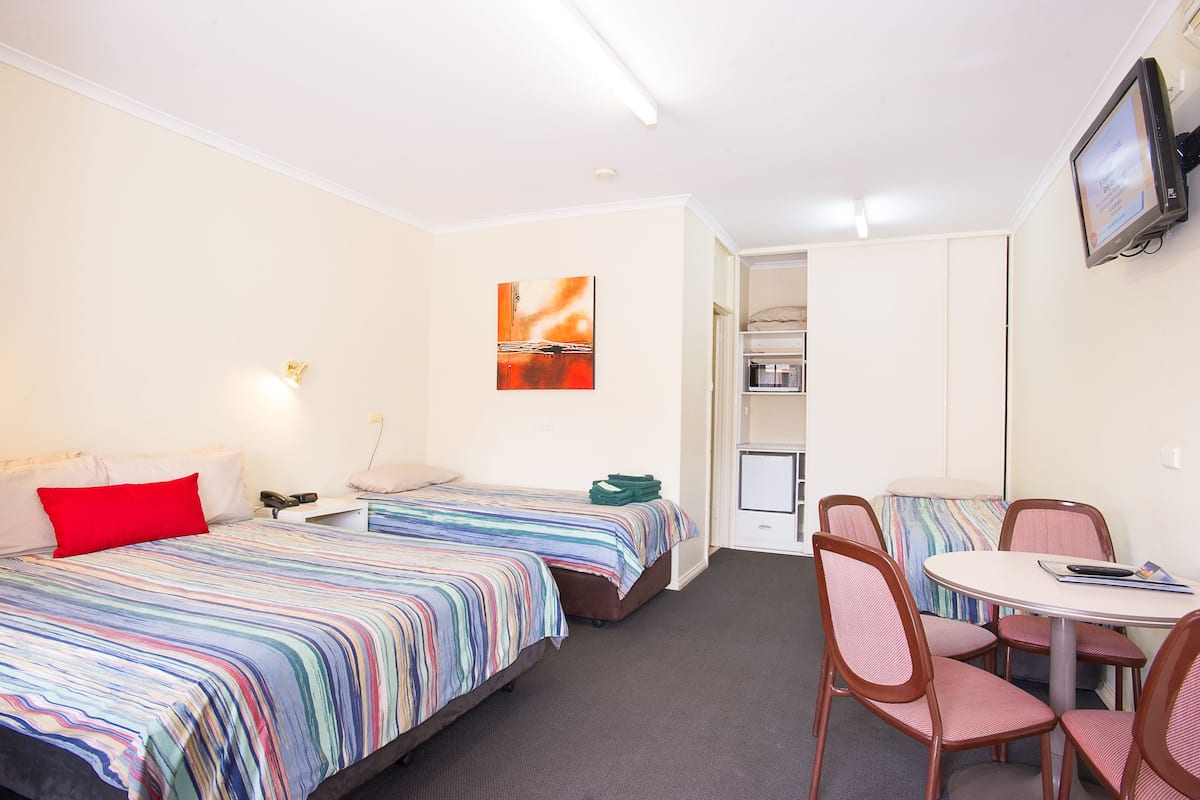 Quarto de hotel com duas camas, uma mesa pequena para refeições e televisão fixada na parede.