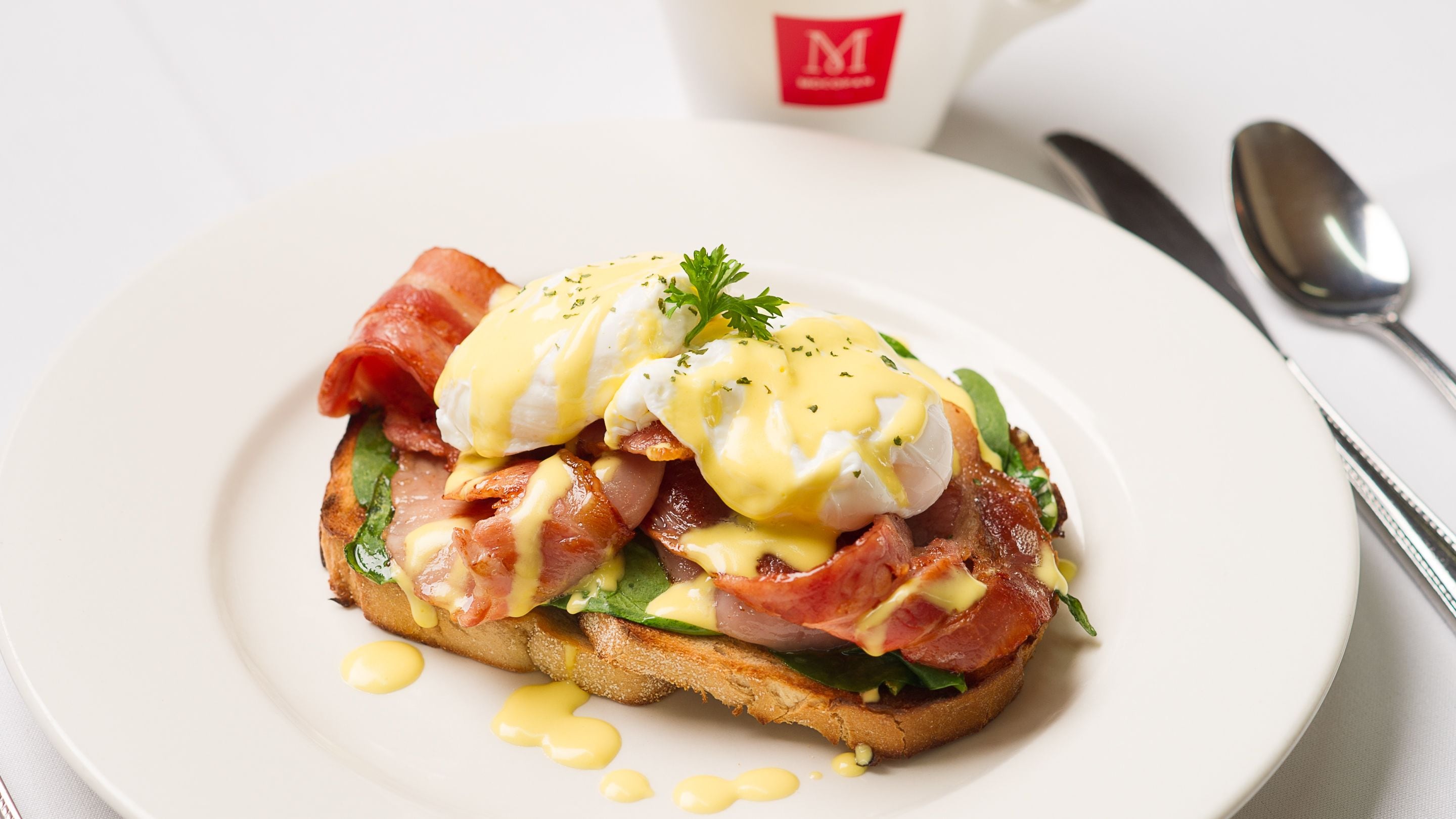 A plate with a breakfast dish, including eggs, bacon, and greens, on a white plate with a spoon and a cup in the background.
