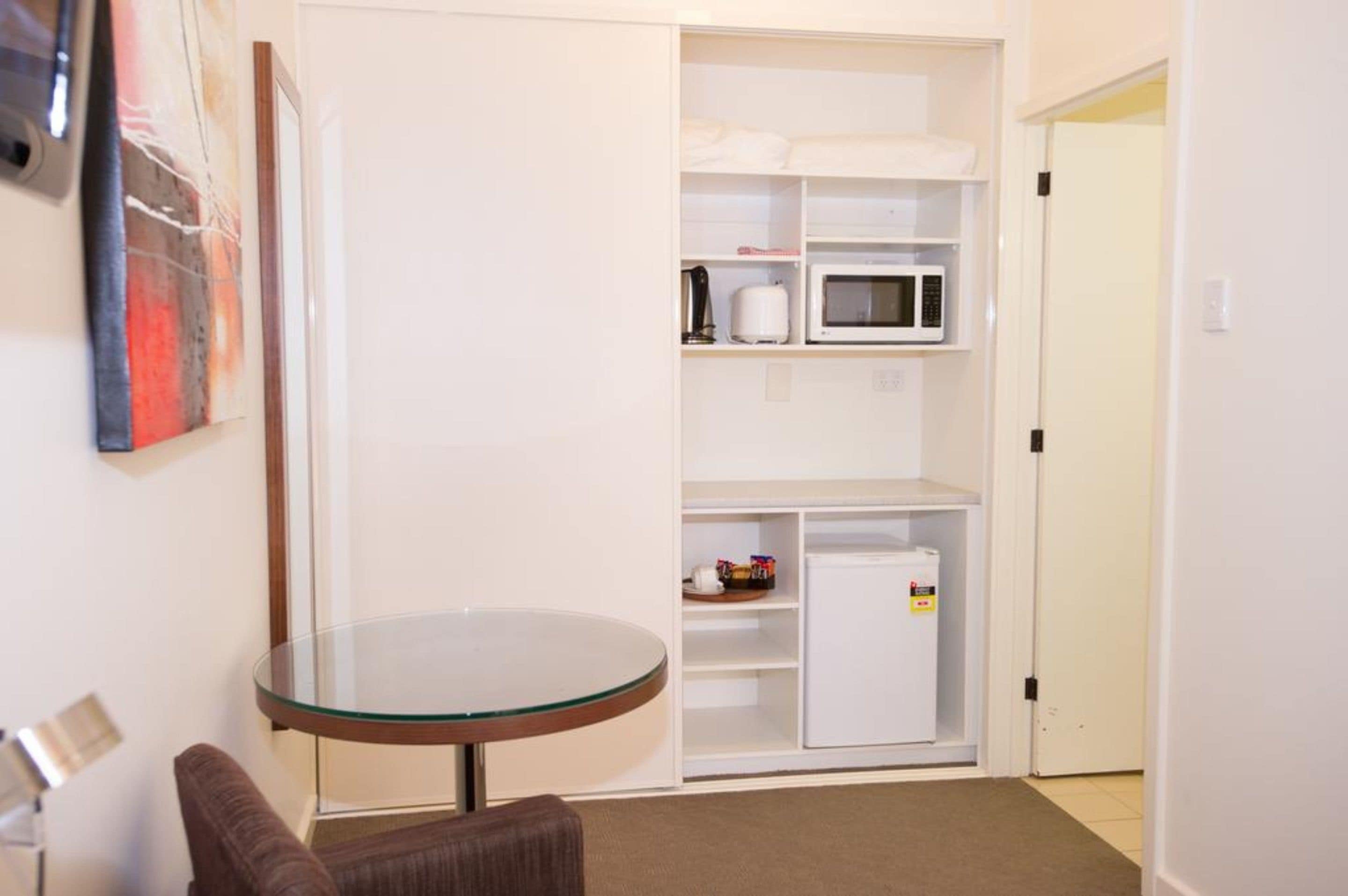 A compact living space with a glass-top table, a sofa, a white wardrobe with shelves, and a microwave.