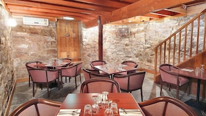 Un coin repas rustique avec des murs en pierre, des poutres en bois au plafond et un escalier. Les tables sont dressées avec des verres à vin et des couverts.