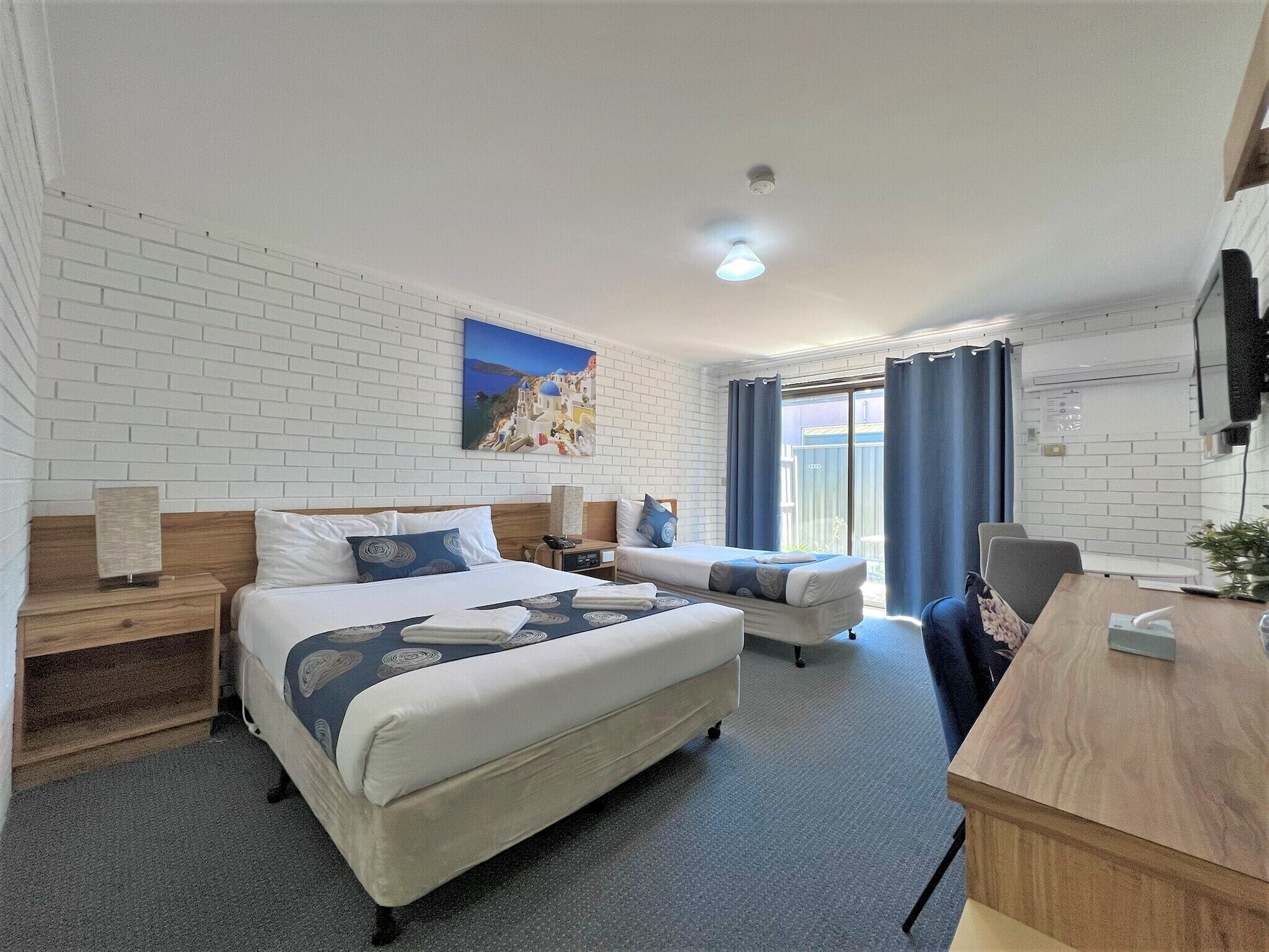 Habitación de hotel con una cama grande, una cama más pequeña, un escritorio y una silla. Hay un cuadro en la pared y una ventana con cortinas.