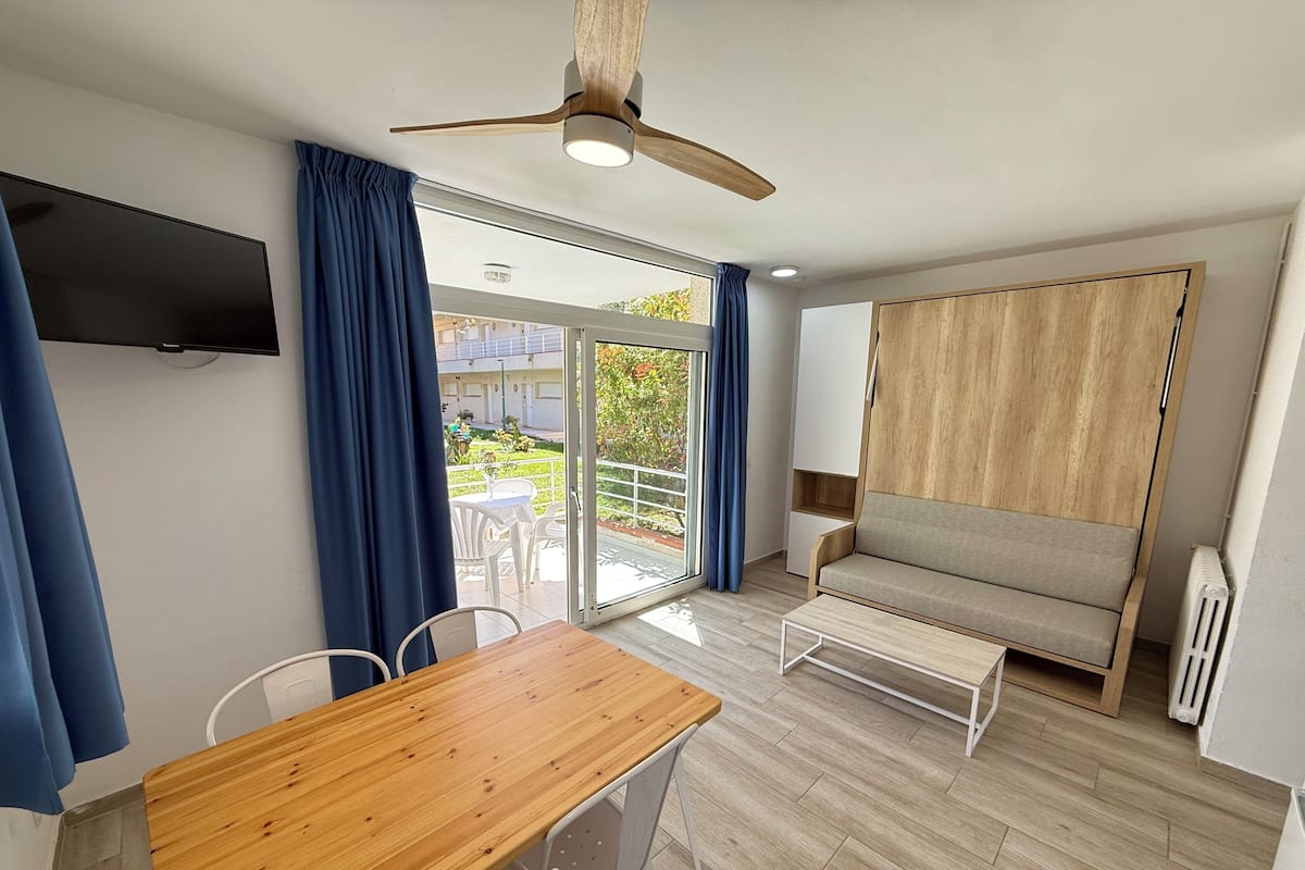 Una habitación moderna con una mesa de madera, sillas blancas, un sofá, un ventilador de techo y un televisor colgado en la pared.