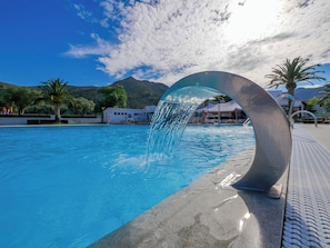 A swimming pool with a water feature, surrounded by palm trees and buildings, under a clear sky.