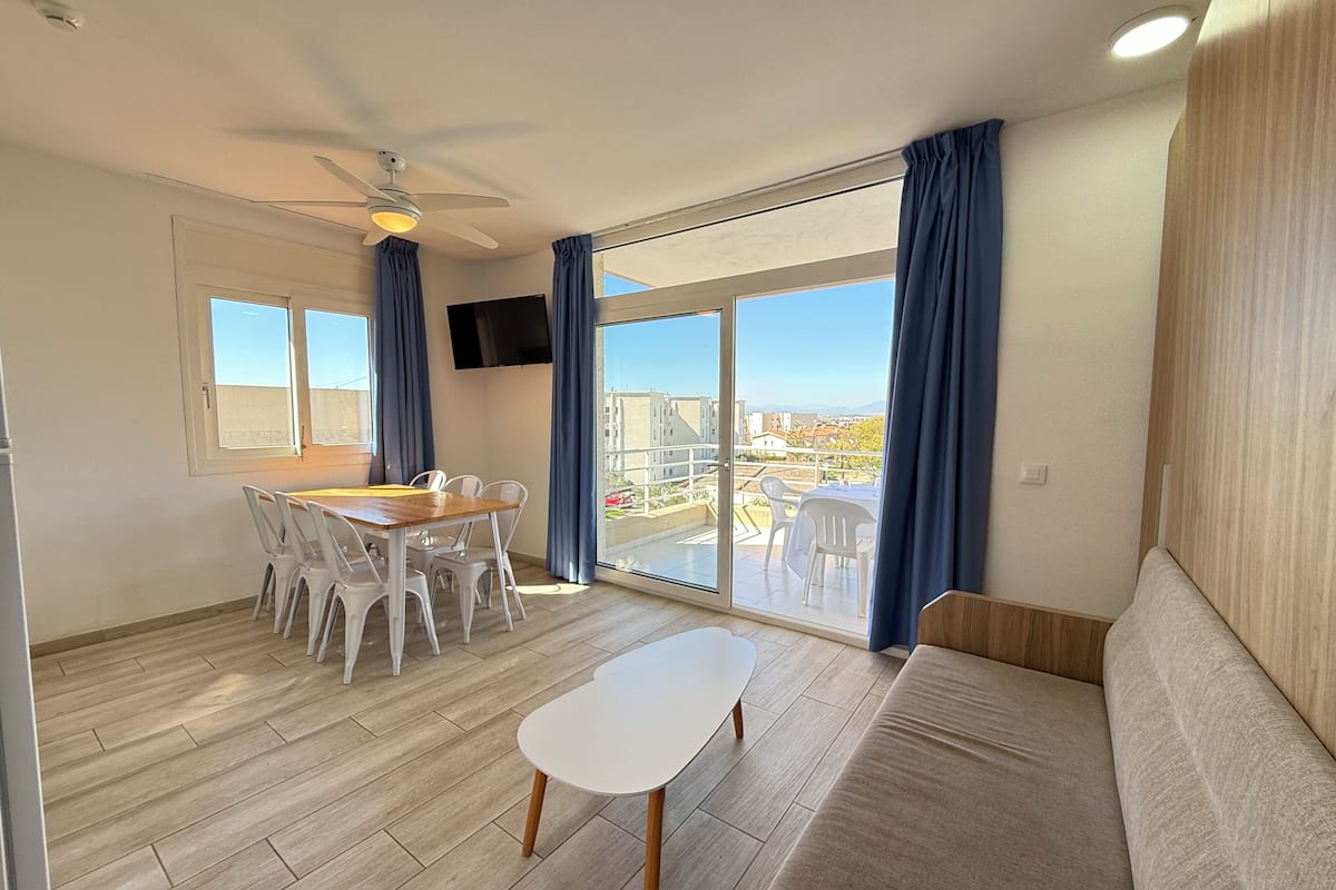 Una habitación moderna con una mesa de comedor, sillas, un sofá, un ventilador de techo, un televisor y un amplio ventanal corredera con vistas a edificios.