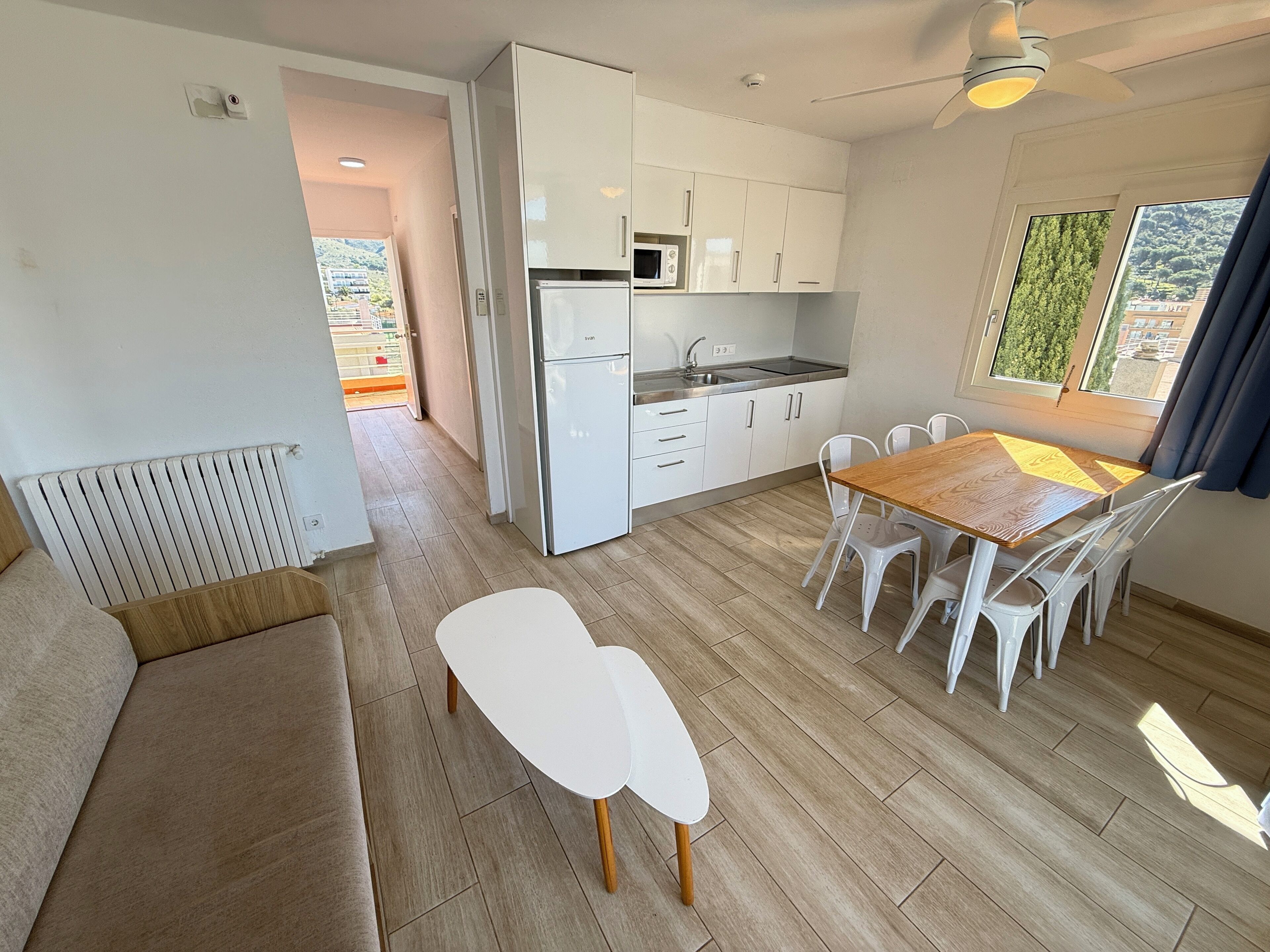 A modern kitchen with white cabinets, a dining area with a wooden table and white chairs, and a view of greenery outside.
