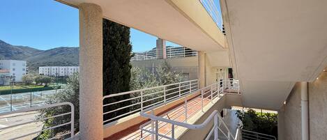 Un balcon extérieur à plusieurs niveaux, doté de rampes de protection, d’un sol carrelé et offrant une vue sur un terrain de sport et des bâtiments en arrière-plan.