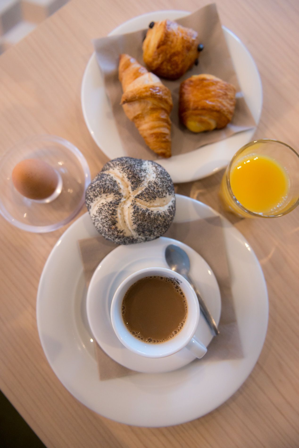 Un desayuno con croissants, un bollo de semillas de amapola, un vaso de jugo de naranja y una taza de café sobre un plato blanco.