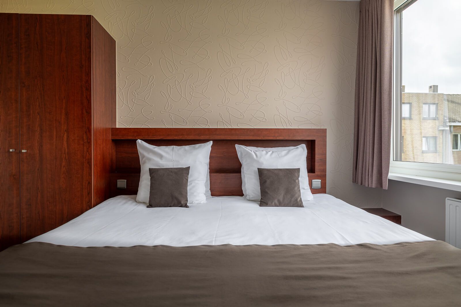 Ein Hotelzimmer mit einem großen Bett, zwei Kissen, einem Holz-Kopfteil und einem Fenster mit Vorhängen.
