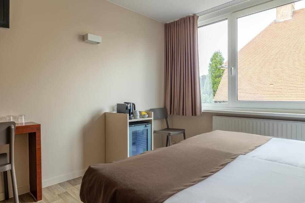 A hotel room with a bed, a desk, a chair, a small kitchenette, and a window with curtains.