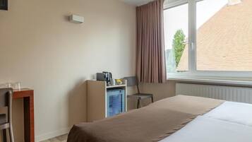 A hotel room with a bed, a desk, a chair, a small kitchenette, and a window with curtains.