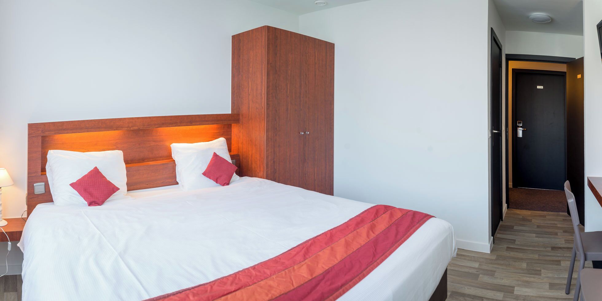 A hotel room with a bed, a wooden wardrobe, a desk, and a door.
