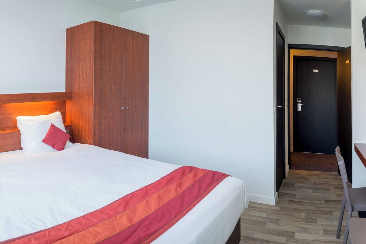 A hotel room with a bed, a wooden wardrobe, a desk, and a door.
